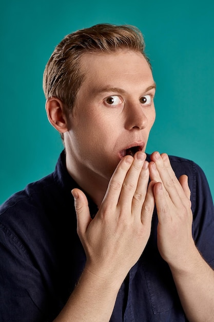 Close-up portrait of a young interesting ginger fellow in a stylish navy t-shirt looking at the camera and acting like he is shocked while posing on blue studio background. Human facial expressions. S