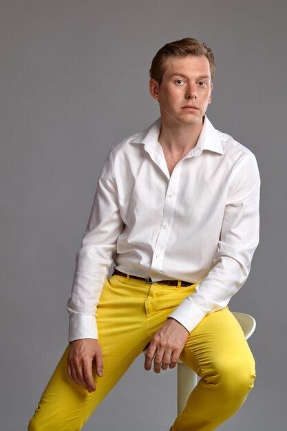 Free photo close-up portrait of a young lovely ginger fellow in a stylish white shirt and yellow trousers looking at the camera and posing sitting on a bar chair against a gray studio background. human facial ex