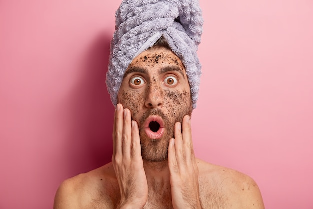 Foto gratuita close up ritratto di giovane uomo pulisce il viso con scrub, guarda con occhi chiusi e bocca aperta, si dimentica della crema cosmetica, indossa un asciugamano, ha il corpo nudo