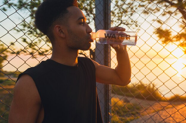 Close-up portrait of young man doing sports in morning, drinking water on basketball court on sunrise