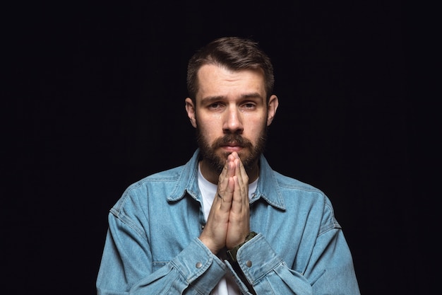 Close up portrait of young man isolated on black  wall. Photoshot of real emotions of male model. Praying and crying, looks sad and hopeful. Facial expression, human emotions concept.