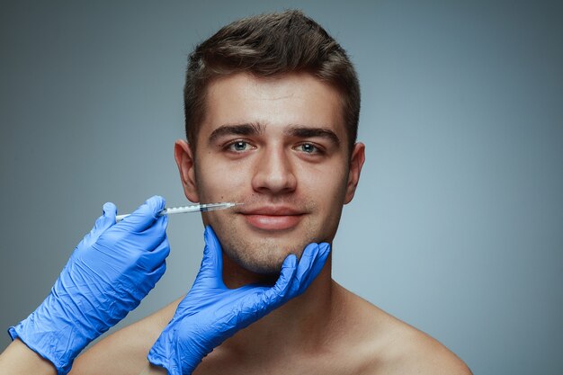 Close-up portrait of young man isolated on grey studio background. Filling surgery procedure. Concept of men's health and beauty, cosmetology, self-care, body and skin care. Anti-aging.