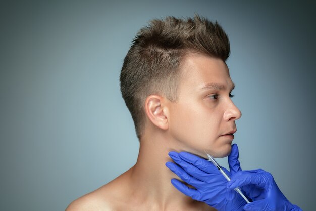 Close-up portrait of young man isolated on grey  wall. Filling surgery procedure, lips and cheekbones. Concept of men's health and beauty, cosmetology, body and skin care. Anti-aging.