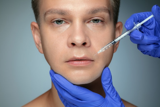 Close-up portrait of young man isolated on grey  wall. Filling surgery procedure, lips and cheekbones. Concept of men's health and beauty, cosmetology, body and skin care. Anti-aging.