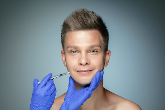 Close-up portrait of young man isolated on grey  wall. Filling surgery procedure, lips and cheekbones. Concept of men's health and beauty, cosmetology, body and skin care. Anti-aging.