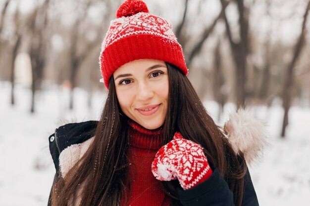 Close up portrait of young pretty candid smiling happy woman in red mittens and knitted hat wearing coat walking playing in park in snow, warm clothes, having fun