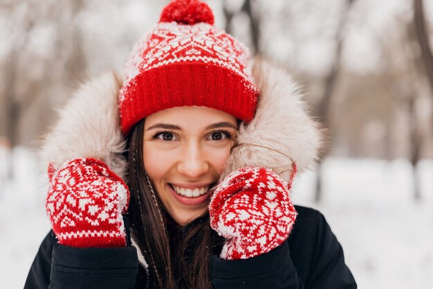 赤いミトンとニット帽を身に着けている若いかなり率直な笑顔の幸せな女性の肖像画をクローズアップ雪の中で公園で遊んで、暖かい服を着て、楽しんで歩いて毛皮のコートを着て