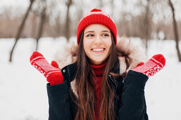 Крупным планом портрет молодой довольно улыбающейся счастливой женщины в красных рукавицах и вязаной шапке в зимнем пальто, прогулки в парке в снегу, теплая одежда