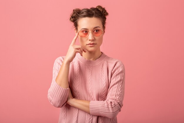 Close up portrait of young pretty thinking woman in pink sweater and sunglasses isolated on pink studio background