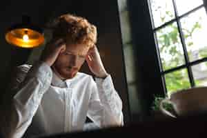 Free photo close-up portrait of young redhead bearded overworked man in white shirt touching his head while sitting at office