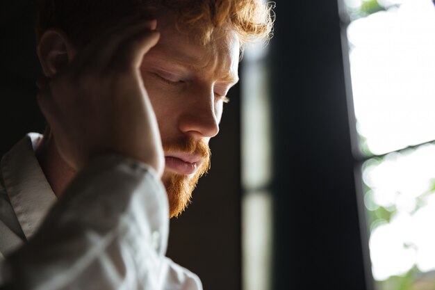 Close-up portrait of young redhead man with headache, touching his head