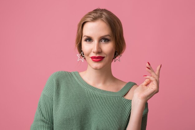 Close-up portrait of young sexy attractive woman, stylish make-up, red lips, green sweater, model posing in studio, isolated, pink background, earrings, looking in camera, holding hand up, elegant