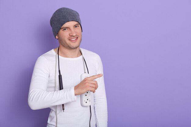 Close up portrait of young smiling electrician wearing gray cap and white long sleeve shirt, holds electric extender on his shoulders and points aside with fore finger. Copy space.