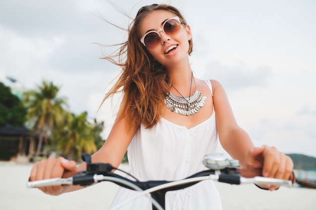 Крупным планом портрет молодой улыбающейся женщины в белом платье, езда на тропическом пляже на велосипедных солнцезащитных очках, путешествующих на летние каникулы в Таиланде