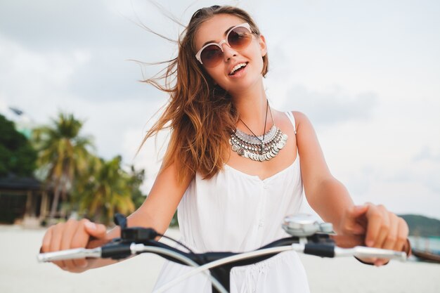 Close up portrait of young smiling woman in white dress riding on tropical beach on bicycle sunglasses traveling on summer vacation in Thailand