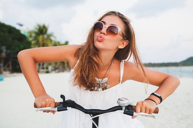 Close up portrait of young smiling woman in white dress riding on tropical beach on bicycle sunglasses traveling on summer vacation in Thailand