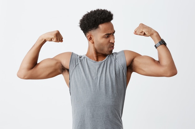 Free photo close up portrait of young sporty dark-skinned man with afro hairstyle in grey shirt showing his muscles, looking at it with concentrated face expression. health and beauty
