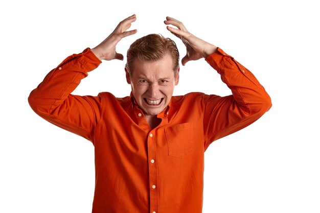 Close-up portrait of a young stately ginger guy in a stylish orange shirt acting like he is beside himself with rage while posing isolated on white studio background. Human facial expressions. Sincere