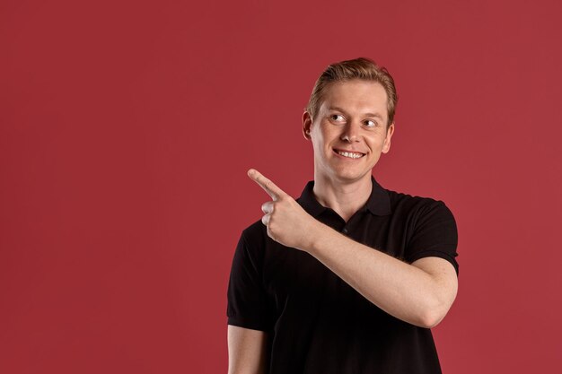 Close-up portrait of a young stately ginger male in a stylish black t-shirt smiling and pointing at something while posing on pink studio background. Human facial expressions. Sincere emotions concept