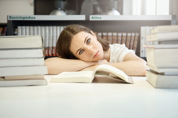 Close up portrait of young student at library preparing for last exams, resting her head on hands, looking sad and tired