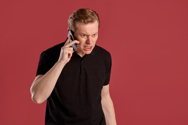 Крупным планом портрет молодого стильного рыжего человека в стильной черной футболке, выглядящего сердитым и разговаривающего по смартфону, позируя на розовом студийном фоне. Выражение лица человека. искренние эмоции