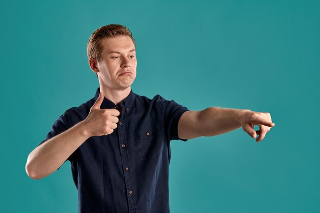 Close-up portrait of a young stylish ginger peson in a navy t-shirt pointing at someone and showing thumb up while posing on blue studio background. Human facial expressions. Sincere emotions concept.