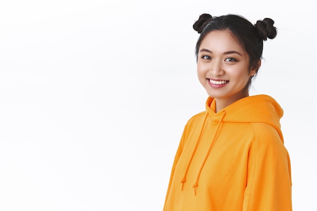 Close-up portrait young teenage asian girl with cute two hairbuns, wear orange hoodie, standing half-turned against white wall near blank empty space for advertisement