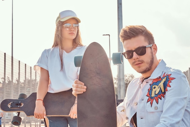 Close-up portrait of young trendy dressed hipster couple with skateboards posing at city sports complex on sunny day, with warm toned.