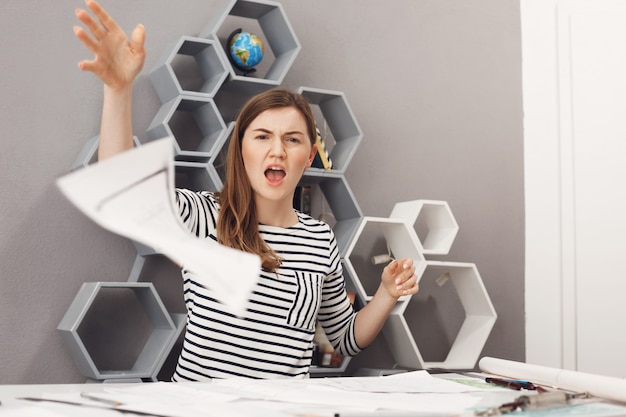 Close up portrait of young unhappy good-looking female freelance engineer throwing work papers away with angry and unsatisfied expression, after customer declined all work.