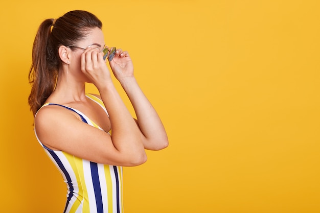 Close up portrait of young woman dressed swimsuit with stripes, holds sunglasses with both hands, posing isolated on yellow