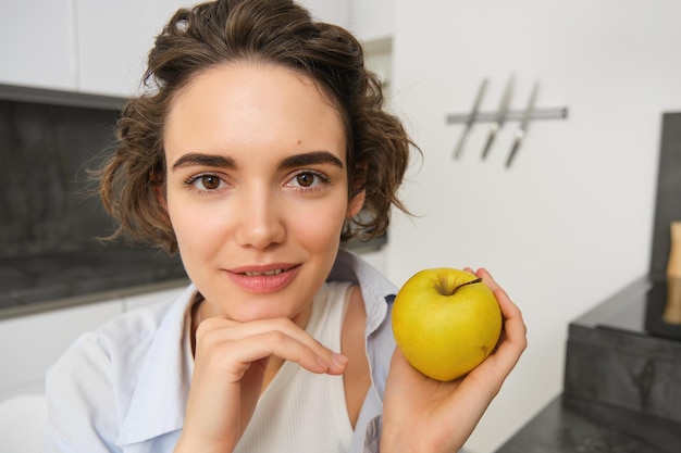 キッチンで幸せに笑顔で健康的な果物を食べているリンゴを握っている若い女性のクローズアップ肖像画