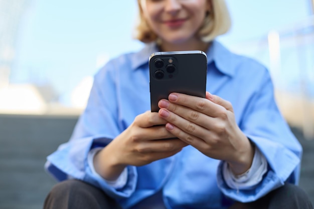 Foto gratuita ritratto ravvicinato di una giovane donna che tiene lo smartphone in entrambe le mani seduta per strada con il cellulare