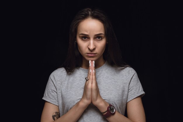 Close up portrait of young woman isolated on black studio background. Photoshot of real emotions of female model. Praying looking forward, sad and hopeful. Facial expression, human emotions concept.