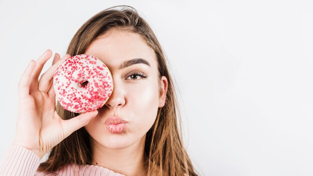 Close-up portrait of young woman pouting lips and covering eyes with donuts isolated on white backdrop