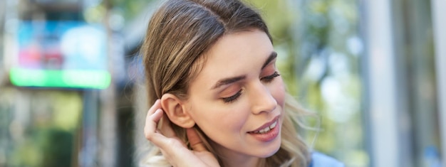 Free photo close up portrait of young woman tuck hair behind ear looking flirty and smiling sitting in blue