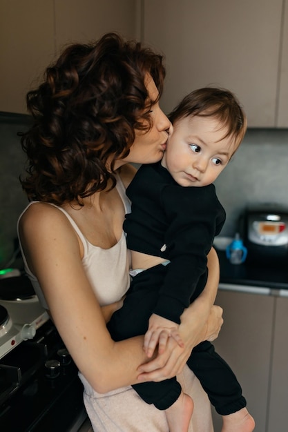 Close up portrait of young woman with wavy hairstyle is wearing home clothes is holding her little son and kissing him home portrait of young woman with her son