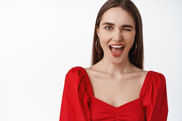 Close up of positive brunette girl in red dress, smiling, winking and showing tongue carefree, being natural, shows true emotions, white background