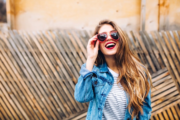 Foto gratuita posrtait del primo piano del magnifico modello femminile in giacca di jeans retrò, tenendo gli occhiali da sole e alzando lo sguardo. sensuale giovane donna con bei capelli lunghi in posa volentieri davanti all'insolita staccionata in legno.