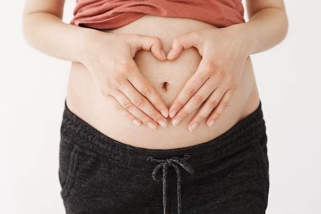 Free photo close up of pregnant belly. young happy mother in casual clothes holds hand on pregnant stomach making heart sign.