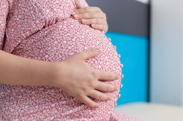Close up of pregnant woman holding hands on baby bump in medical office. patient with pregnancy expecting child and preparing to give birth. person with baby in womb at checkup visit