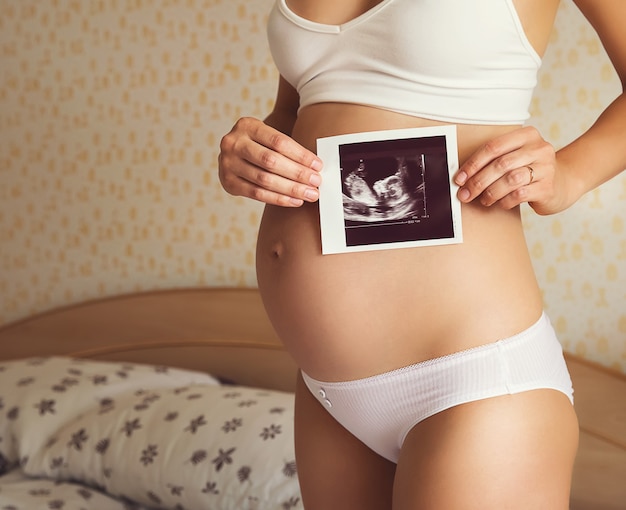 Close-up pregnant woman holding ultrasound image in home interior.