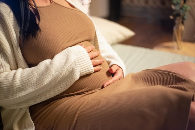 Close-up of pregnant woman sitting on bed. Woman in dress and cardigan touching big belly. Pregnancy, expectation concept