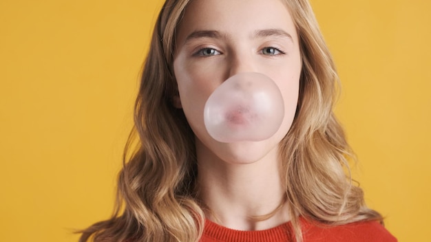 Close up pretty blond teenager girl with wavy hair blowing bubble from gum on camera over yellow background. Candy bubble