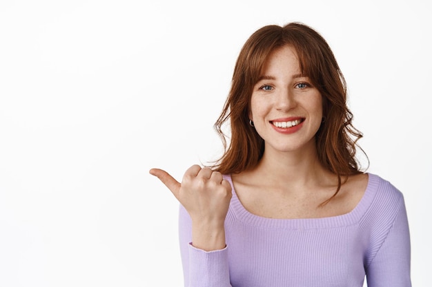 Primo piano di bella ragazza con frangetta, nuova acconciatura e trucco sul viso, puntando il pollice a sinistra, sorridente felice con denti bianchi perfetti, in piedi in camicetta su sfondo bianco.