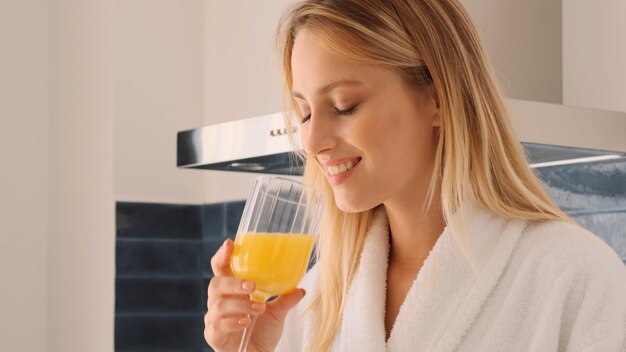 Close up pretty smiling blond girl in white bathrobe happily drinking orange juice on kitchen at home Attractive girl having breakfast