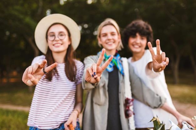 Free photo close up pretty smiling girls happily showing victory gestures and looking in camera in park