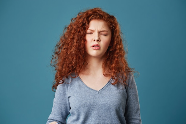 Close up of pretty young woman with wavy ginger hair and freckles in grey shirt having headache after sleepless night.