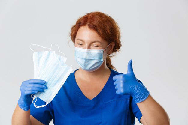 プロの女性看護師のクローズアップ、医師はウイルスの発生を防ぐためにフェイスマスクと保護装置を着用することをお勧めします、親指を立てる