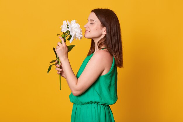 Close up profile portrait of lady wearing green sundress keeps flowers in hands on yellow, being happy to recive peonies as gift.