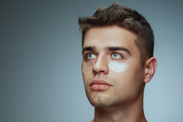 Close-up profile portrait of young man isolated on grey studio background. Male face with collagen patches under eyes. Concept of men's health and beauty, cosmetology, body and skin care. Anti-aging.